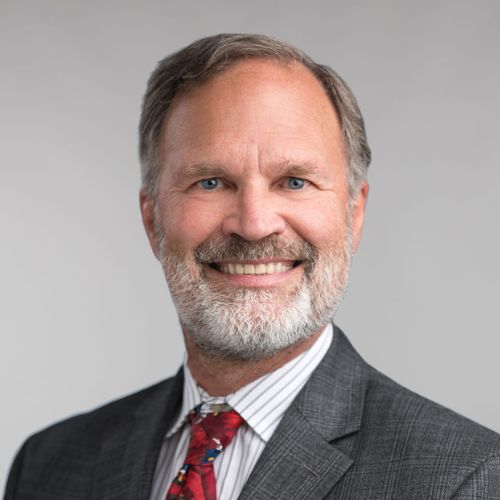 Smiling middle-aged man with gray hair and beard, wearing a gray suit, white striped shirt, and red patterned tie, posed against a plain light gray background.
