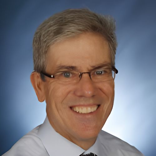 Smiling middle-aged man with short gray hair, glasses, and a white shirt with a tie, posing in front of a softly blurred blue background.