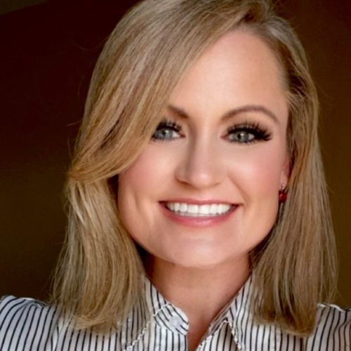 A woman with shoulder-length blonde hair smiles at the camera. She is wearing a white blouse with black vertical stripes and has bright makeup, including eyeliner and lipstick. The background is a plain, warm brown color.